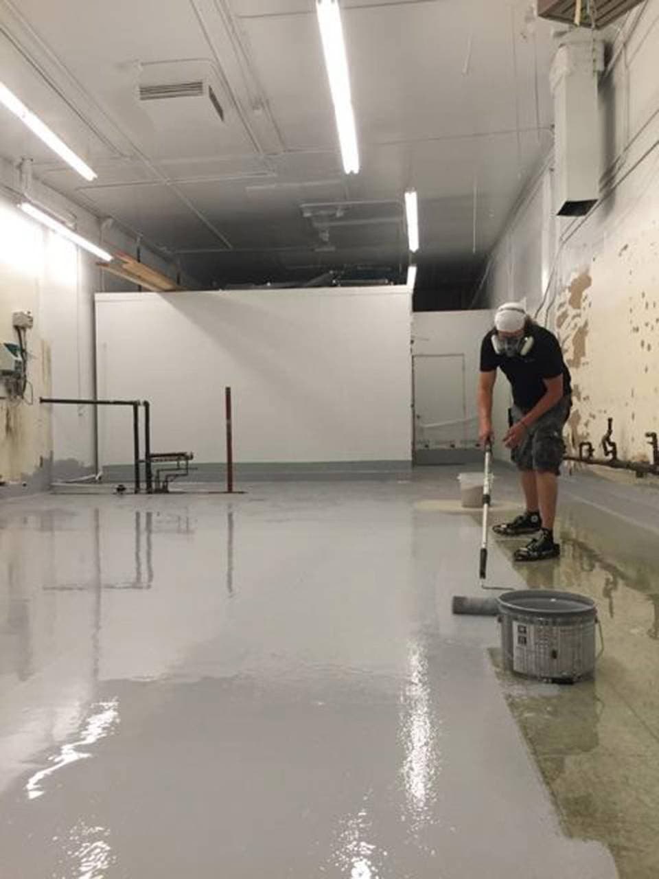 A worker in a black shirt, shorts, and a white cap is applying a coat of paint or epoxy on a concrete floor inside a commercial space, with a bucket and roller nearby, suggesting ongoing floor coating work for a painting business.