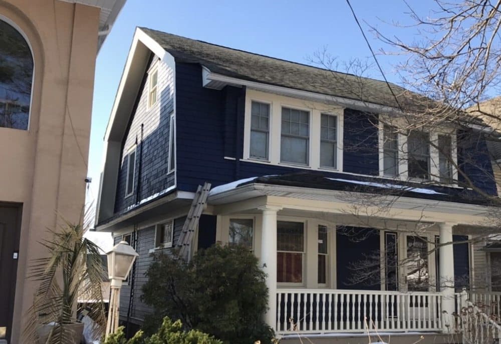 A two-story house with a dark blue exterior, white trim, and a covered front porch supported by white columns, featuring a ladder leaning against the side, indicating recent painting or epoxy floor coating work.