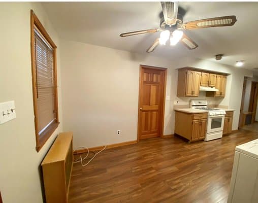 A kitchen with wooden cabinetry, a white stove, and laminate flooring, illuminated by a ceiling fan with lights, with a window and a wooden door on the walls.